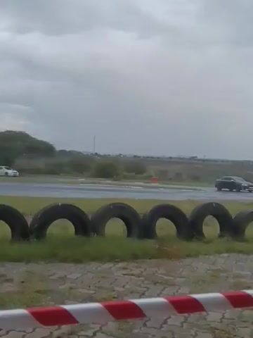 The video shows two cars driving on a wet racetrack on an overcast day. In the foreground, a row of tires separates the viewer from the track. A white car is seen passing first, followed by a gray car with its headlights on. The cars are driving around a curve in the track, and the surrounding area consists of grassy fields and some scattered trees. In the distance, buildings can be seen on the horizon under a cloudy sky.