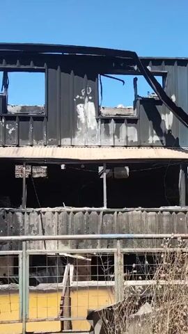 The video shows the aftermath of a fire at a two-story building made from shipping containers. The building is heavily damaged, with windows broken and the exterior charred. A metal beam is hanging down from the roof of one of the containers. There is debris scattered around the base of the building. Several people are walking around the scene, including one person climbing the metal staircase. The sky is clear and blue.