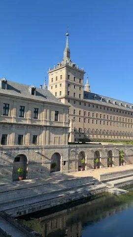 The video pans across a large, ornate building with a central tower topped by a spire. The building is made of stone and has many windows. In front of the building is a body of water, possibly a pond or canal, with a stone walkway alongside it. The water reflects the sky and the building. Beyond the building and walkway, there is a landscape with trees and rolling hills under a clear blue sky. The scene appears to be a historical site or a grand estate. There are no people visible in the video. The lighting suggests it is daytime.