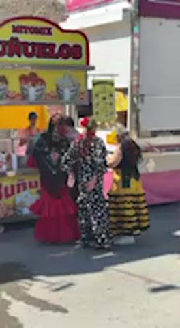 This video captures an outdoor scene, likely a fair or festival, in Jerez de la Frontera, Spain, on a sunny day. Several people are gathered near a yellow food stall. The stall features the prominent text "BUÑUELOS" and displays various food items in buckets.

Among the people, three individuals are notably dressed in traditional Spanish attire. One person wears a striking black and white polka-dotted flamenco-style dress, another is in a vibrant red dress with ruffled tiers, and a third wears a yellow and black striped dress. These individuals appear to be dancing or engaging in some form of rhythmic movement, with their arms raised and skirts swaying. A man in a light-colored jacket and dark pants stands to the left, facing the food stall. The ground is paved, and strong shadows suggest bright sunlight.
