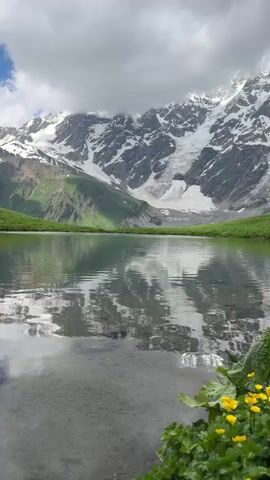A still shot of a mountain lake with the reflection of snow-capped mountains and clouds on its surface. The foreground features green grass and yellow wildflowers.