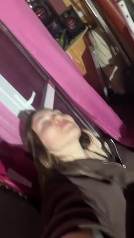 The video shows a young woman lying down with her eyes closed. She is wearing a brown zip-up hoodie. There is a pink fabric to her left. The lighting is dim, and the background is out of focus, suggesting it may be nighttime.