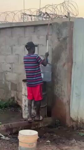 A young male stands atop a rusted metal barrel, dressed in a blue-and-red striped shirt, red shorts, and a black baseball cap. He is positioned next to a weathered concrete block wall topped with barbed wire. He grips a long, white PVC pipe attached to a large, clear plastic water jug. The jug appears to be filling with water from the pipe. The environment consists of dirt and minimal vegetation, with hints of an industrial or potentially hazardous area. A white bucket sits on the ground nearby. The overall lighting suggests it is either early morning or late afternoon.