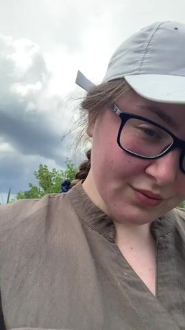 A young woman with brown hair pulled back in a braid and wearing glasses and a white baseball cap is taking a selfie outdoors. She is wearing a khaki-colored shirt. In the background, there are green trees and a body of water, possibly a river, with a forest on the opposite bank. The sky is cloudy. The woman smiles and tilts her head slightly in the first few frames, then turns to show more of the scenery in the last frame.