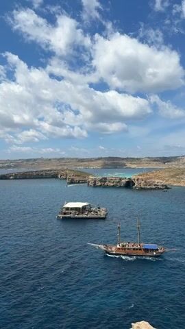 The video shows a seascape with a rocky, arid coastline in the background, a bright blue sea, and a partly cloudy sky. In the foreground, there is a wooden sailboat with people on board, sailing from right to left. Further away, there is a flat, rectangular structure with a white roof floating in the water. The water is calm and blue, with some areas appearing more turquoise. The coastline in the background is mostly brown and rocky, with some green vegetation. The sky is blue with fluffy white clouds.