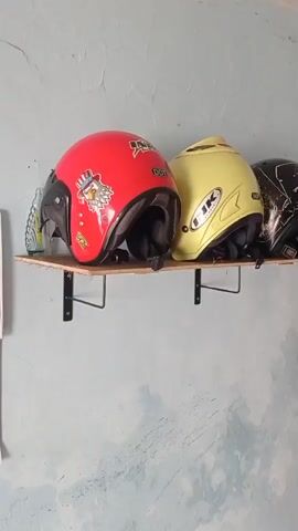 The video shows a close-up shot of three motorcycle helmets displayed on a wooden shelf mounted on a pale blue, textured wall. The leftmost helmet is red and features a sticker of an eagle wearing a hat. Next to it is a yellow helmet with a black logo that appears to be "INK". The third helmet, partially visible on the right, is black with a white abstract pattern. The shelf is supported by two black L-shaped brackets. To the right of the helmets, a dark fabric item, possibly a jacket or coat, is hanging on the wall, partially obscuring the view of the wall and a pink curtain. The lighting appears to be natural, coming from an unseen source, casting soft shadows. The overall scene suggests a storage area or a display of protective gear.