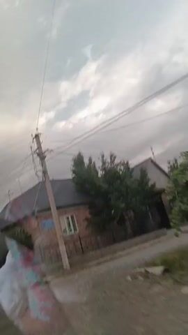 The video shows a moving shot from inside a vehicle, looking out at a cloudy sky, telephone wires, and buildings. A brick house with a dark roof is visible on the left, with a blue sign on the wall. Trees and a fence are also present. Further down the street, another building with a green and white section and a red sign is seen. A white car is parked in front of it. The scene appears to be a residential or semi-commercial area.