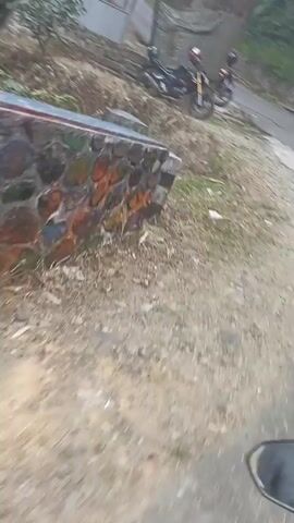 The video opens with a view of a dirt road and a motorcycle parked on the side. Behind the motorcycle is a structure with a corrugated metal roof and a stone wall. Trees and vegetation are visible in the background. The camera then pans to the right, revealing a tree covered with a checkered cloth. The video continues to pan, showing more of the surrounding environment, including more vegetation and what appears to be a small building or shed. The overall scene suggests a rural or semi-rural setting.