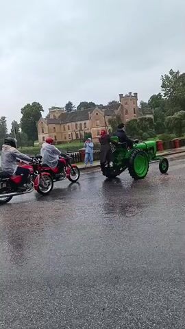 The video shows a procession of several vintage motorcycles and a green tractor driving on a wet road. In the background, there is a large, light-colored building that appears to be a castle or manor house, surrounded by trees. The sky is overcast and it is raining. The motorcycles are moving from right to left in the frame, and the tractor is also moving in the same direction. Some of the motorcycles have riders wearing helmets and rain gear. There are also a few people standing on the side of the road watching the procession. One of the motorcycles has a sidecar. The road is wet and reflects the overcast sky. The overall scene appears to be a parade or rally in wet weather.