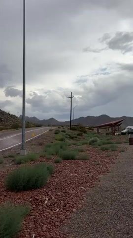 This is a video of a highway in a desert environment. The sky is overcast with dark clouds, suggesting the possibility of rain. The highway is paved with a double yellow line in the center and a white line on the side. The landscape is characterized by rocky hills covered in sparse desert vegetation, including shrubs and some cacti. There are utility poles and streetlights visible along the road. A curve warning sign is also present. The ground along the roadside is covered in small reddish-brown gravel with patches of dry grass. The scene appears to be uninhabited.