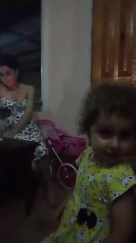 The video shows a young child with curly hair wearing a yellow outfit with cartoon characters. The child is in the foreground and appears to be looking at the camera. In the background, an older man with gray hair is seated and looking to the side. To the left, a woman in a patterned dress is sitting and looking at her phone. A pink toy stroller is visible near the woman. The environment appears to be indoors, possibly a living room, with wooden doors in the background. The lighting is dim.