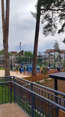 The video captures an outdoor sports and recreation area on a cloudy day. In the foreground, a wooden deck with gray railings offers a raised vantage point. The view extends to a fenced sports court with a net, possibly for volleyball or tennis. Several people, including adults and children, are scattered throughout the court and on a nearby playground with blue equipment. Some individuals appear to be exercising on outdoor fitness apparatus located to the left of the sports court. The area is surrounded by trees, some of which are tall pines, and residential buildings can be seen in the background. The overall impression is of a community space being utilized for recreational activities.