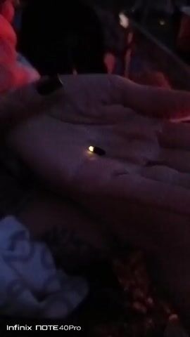A hand is held out, palm up, with a single firefly resting on it. The firefly is small and black, with a bright yellow light glowing from its abdomen. The background is dark and out of focus, with some blurry red and purple lights visible. The image is captured with an Infinix NOTE 40Pro phone.