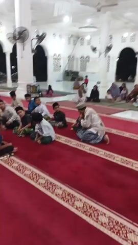The video shows a group of young boys sitting on a red carpet inside a mosque. They are facing a man who appears to be teaching them. The man is wearing traditional Islamic clothing. Some of the boys are wearing similar clothing, while others are wearing t-shirts and shorts. The boys are sitting in rows, and they are all facing the same direction. There are also some women and children sitting in the background, but they are not as clearly visible. The mosque is decorated with white columns and arches, and there are fans on the ceiling to keep the people cool. The lighting in the mosque is bright and even. The video appears to be filmed from a low angle, looking up at the group of boys and the teacher.