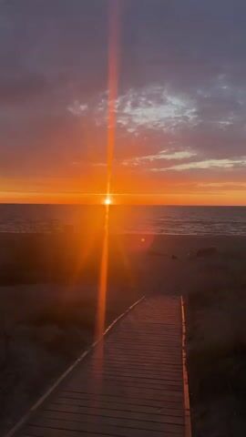 The video shows a sunset over a beach. A wooden boardwalk leads towards the beach and the ocean. The sun is a bright orange and casts a beam of light down towards the boardwalk. The sky is filled with clouds, colored in shades of orange, pink, and gray. The water is calm. There are patches of beach grass on either side of the boardwalk. A voice speaks in the background but is unintelligible.