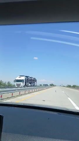 The video shows a view from inside a car driving on a highway. The sky is clear and blue with a few white clouds. On the left side of the road, there is a car carrier truck loaded with several vehicles. The highway has multiple lanes and a concrete barrier separating it from the other side. In the distance, other vehicles can be seen on the road. The interior of the car, including the dashboard and windshield, is visible in the foreground. The overall scene depicts a typical highway travel experience on a sunny day.