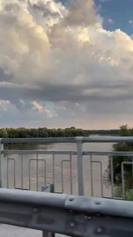 The media file captures a scenic view from a bridge overlooking a wide, calm river. The river, appearing muddy brown, flows gently between lush green banks lined with dense trees. A silver metal railing runs along the foreground, with a patterned design of vertical bars and horizontal rectangles, suggesting the perspective is from a moving vehicle on the bridge. In the distance, a faint rainbow arches across the sky, hinting at recent or impending rain.

The sky is dramatic, with a mix of dark, brooding storm clouds and patches of bright, fluffy white clouds illuminated by the setting sun. The light suggests it is either late afternoon or early evening, with a warm, golden hue cast over the landscape. The weather appears to be transitioning, with the possibility of a storm passing.

There are no people visible in the media file. The primary subjects are the natural landscape of the river and sky, and the man-made structure of the bridge railing. The scene evokes a sense of calm and natural beauty, with a touch of drama from the dynamic sky.

A white rectangular sign is partially visible on the left side of the bridge, attached to the railing structure. It appears to have some text or symbols on it, but they are not clearly legible. The location context provided is Ladozhskaya, Russia, which places the scene in a rural or semi-rural setting with a significant river.
