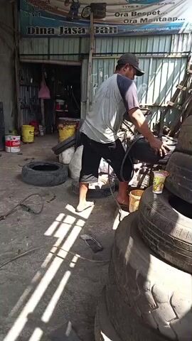 The video shows a man in a garage or workshop wearing a cap and a t-shirt, working on a stack of tires. He is holding a hose and appears to be cleaning or inspecting the tires. The environment is cluttered with tires, tools, and other equipment. The lighting suggests it is daytime, with sunlight streaming into the space, creating shadows. There are signs on the wall, one of which reads "Dina Jasa Ban" which translates to "Dina Tire Service". The man is barefoot, wearing shorts. He is actively engaged with one of the tires, suggesting a process of tire repair or maintenance. The overall scene depicts a busy, functional workshop environment.
