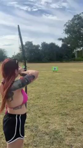 A woman with pink hair and tattoos on her left arm and torso is practicing archery. She is wearing a pink bikini top and black shorts with white trim. She is standing in a grassy field, aiming a black bow at a green target with a bullseye painted on it. There are trees in the background, and the sky is partly cloudy. The woman pulls back the bowstring and aims, and then the video ends before she releases the arrow.