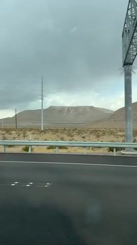 The video shows a desert landscape with a cloudy sky. A highway runs through the foreground, with a dry, dusty terrain stretching out behind it. In the distance, mountains are visible. A large billboard stands on the left side of the road, and power lines stretch across the sky. The video appears to be filmed from a moving vehicle, as the perspective shifts slightly. There are no people visible in the video. The overall impression is one of a vast, arid environment under a heavy sky.