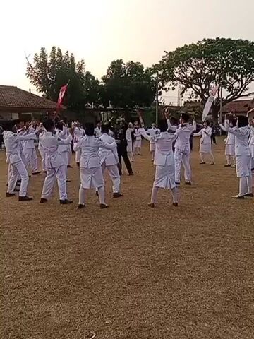A group of individuals dressed in white uniforms are standing in a circle on a dry, grassy field. Some individuals are holding flags or batons. The sky is overcast with the sun setting in the background. There are trees and buildings visible in the distance. The individuals appear to be engaged in a coordinated activity or performance. One individual in a darker uniform is in the center of the circle, seemingly directing the group. The overall atmosphere suggests a formal event or training exercise.