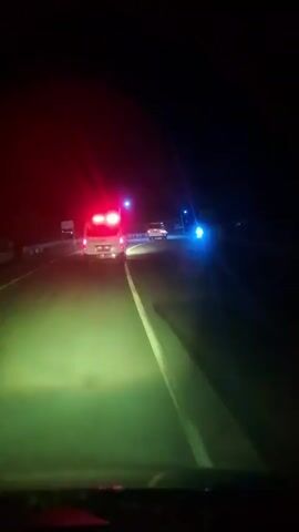 The video shows a first-person view from a car driving on a dark road at night. The car's headlights illuminate the road with a yellow-green light. Ahead, an ambulance with flashing red lights is visible, driving in the same direction. Another vehicle with bright white headlights is behind the ambulance. Streetlights with blue and white lights are seen along the road. The video has sound, including an ambulance siren and music playing in the car.