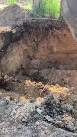 An excavator bucket is digging into a trench. The trench appears to be filled with dirt and debris, and there are tree roots visible along the side of the trench. The excavator is orange and yellow. The background shows grass and a fence.