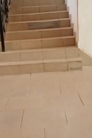 The video shows two women descending a set of tiled stairs. One is wearing a long purple dress and a white hijab, carrying a pink tote bag. The other is wearing a light blue patterned dress with a black jacket, also carrying a pink tote. The stairs have a black metal railing on one side. The walls are painted a light color.