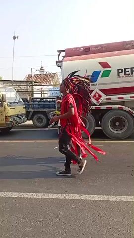 A parade is taking place on a paved road. Several people dressed in red and black traditional outfits are walking and dancing. A large red and white tanker truck with the Pertamina logo is parked on the side of the road, and several other trucks are visible behind it. The sky is bright and clear, suggesting it is daytime. The overall scene depicts a festive and cultural event.