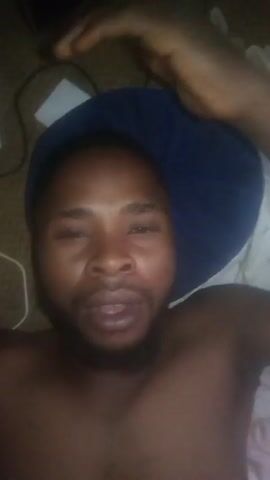 A shirtless man lies supine on a bed with a blue pillow under his head. He alternates between covering his mouth with his hand and smiling or grimacing. A white charging cable and a small white object are visible near his head on the bed. A partially visible white blanket or sheet is in the background. The lighting is dim, and the camera angle is directly above the man's face.