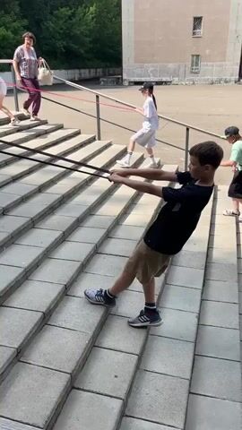 A group of people are engaged in an outdoor activity on a wide set of concrete stairs. A young boy in a black t-shirt and khaki shorts is in the foreground, facing away from the camera, pulling on two thick black resistance bands that extend upwards and to the left. He is standing on the stairs, leaning back with his arms extended. To his left, further up the stairs, a young girl in a white t-shirt and white shorts, wearing a black baseball cap, is also pulling on resistance bands. Behind her, a woman in a patterned blouse and burgundy pants is walking down the stairs, holding a light-colored bag. The environment appears to be a schoolyard or park, with trees and a building visible in the background. The lighting suggests it is daytime.