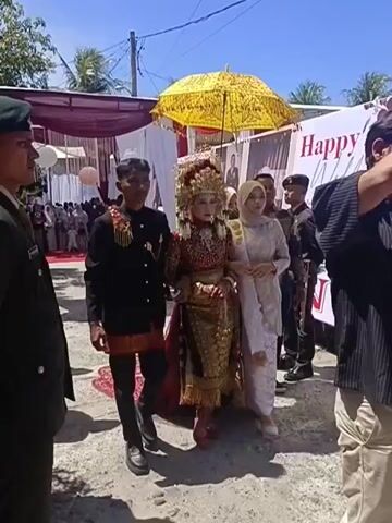 The video shows a wedding procession outdoors on a sunny day. A bride, adorned in a traditional elaborate headdress and a richly decorated red and gold outfit, is walking between two men. The man on her left is wearing a black jacket with red trim and a matching sarong. The man on her right is wearing a light-colored, possibly lace, dress and a hijab. Behind them, a person holds a large, ornate yellow umbrella over the bride. Two men in military uniforms are visible on the left and right sides of the frame, appearing to be part of the procession or security. In the background, a wedding banner reads "Happy Wedding" and other decorations suggest a festive event. People are gathered in the distance, and tents are set up. The ground is unpaved. The bride and her companions are walking towards the camera.