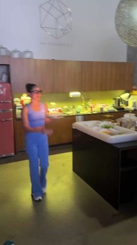 A woman in a blue workout outfit holds a lit birthday cake with candles and walks toward a group of people standing and sitting at a long table. Behind her is a kitchen area with brown cabinets and a pink refrigerator. The people at the table are facing her and singing "Happy Birthday." A red backdrop is visible in the background. Above, there are geometric light fixtures. A white wall says "EM COSMETICS". There is a man standing near the kitchen. The video is somewhat shaky.