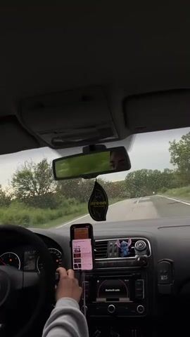 The video, shot from inside the driver's seat of a car, shows a driver navigating a rural road in rainy weather. The windshield wipers are in motion, clearing raindrops from the glass. The rearview mirror reflects the face of the driver, who has dark eyes. In the foreground, the driver's hand rests on a phone mounted in a holder, displaying a music playlist. The car's dashboard is visible, with a small Stitch figurine attached to the air vents. An "Areon" air freshener hangs from the rearview mirror. The car's speedometer reads 25 km/h. The car's radio displays "Bluetooth audio." The road ahead stretches into the distance, flanked by trees. The sky is overcast, adding to the somber atmosphere of the scene.