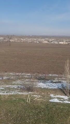The scene depicts a rural landscape on a sunny day. A large, plowed field dominates the view, its brown earth contrasting with patches of melting snow in a lower area. Some grassy areas are visible in the foreground and in the background, with some scrub and leafless trees scattered in the lower area, behind which the plowed field extends into the distance.  A small village is visible on the horizon, with what appears to be a tall structure, perhaps a tower or silo, rising above the buildings. In the far left of the frame, a utility pole stands starkly against the skyline. The sky is pale blue with some wispy clouds. There are no people visible in the video.