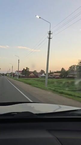 The video shows a view from inside a moving vehicle, looking out the windshield. The vehicle is on a paved road in a rural area. On the right side of the road, there is a grassy field with several houses and fences. Trees are visible in the background. The sky is a pale blue with some clouds, and the sun appears to be setting, casting a warm glow. There are utility poles and power lines along the side of the road. The road is in good condition, with a white line marking the edge. The interior of the vehicle is visible at the bottom of the frame, showing a black dashboard. The video is short, lasting only a few seconds.