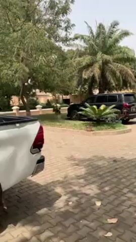 The camera pans across a paved area to reveal several parked vehicles and a motorcycle in a landscaped yard. A white Toyota Hilux pickup truck is visible in the foreground. Further into the frame, there is a black SUV, a red car, and another dark-colored SUV. A red motorcycle is parked on the paved area, and beyond it, a grassy lawn extends to a peach-colored wall. Mature trees and tropical plants, including palm trees, dot the landscape. The overall scene suggests a residential or commercial property with ample parking. The sky is overcast, and the lighting is diffused.