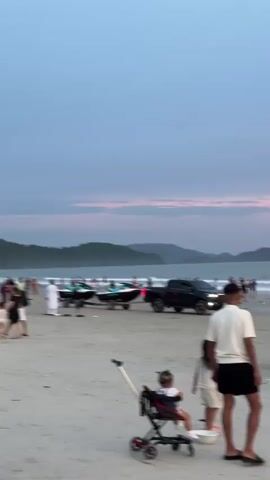 The video shows a beach scene during dusk. Several people are present on the sand, including a man in a white shirt and black shorts bending down, a young child in a stroller, and another child standing nearby. In the background, there are several jet skis on trailers being pulled by a dark-colored truck. More people are visible in the distance, some walking along the shoreline and others in the water. The ocean stretches to the horizon, with islands or hills visible in the far distance. The sky displays a gradient of soft blues and pinks, indicative of sunset.