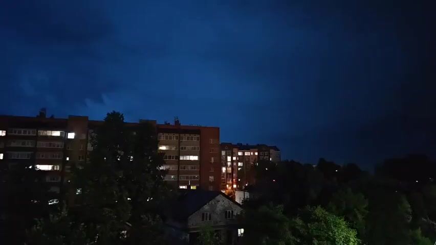 A still shot captures a darkened sky over an urban landscape at dusk or night. The sky is a deep blue, with wisps of clouds visible, hinting at an impending storm or already existing cloud cover. Below the sky, the outlines of several apartment buildings are visible, characterized by multiple stories and windows. Many of these windows are illuminated with a warm, yellowish light, suggesting that the apartments are occupied and that it is nighttime. In the foreground, dark silhouettes of trees obscure the lower portions of the buildings and also contribute to the overall dark ambiance of the scene. The overall impression is one of a quiet, residential area under a moody, darkening sky.