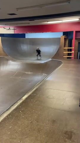 The video captures a person skateboarding in an indoor skatepark. The skatepark features a large half-pipe with wooden ramps and a concrete floor. The person, wearing protective gear including a helmet, knee pads, and elbow pads, is seen riding up and down the ramps, performing tricks. At one point, they launch into the air, executing a mid-air maneuver before landing back on the ramp. The scene is well-lit with overhead lighting, and the surrounding walls of the skatepark are painted with red and blue accents. The camera angle shifts, providing different perspectives of the action. In the final frames, the skateboarder is seen kneeling on the ramp, with their skateboard nearby, suggesting a pause or the end of a trick.
