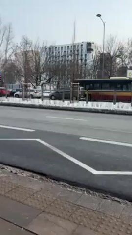 The camera is static, positioned on a sidewalk overlooking a wide city street with several lanes of traffic. The asphalt road surface is clean and dry. A bus is parked along the side of the road, its doors apparently open. Several cars are parked behind the bus. A modern building is visible in the background. The sky is pale blue. There is some snow on the ground alongside the road, next to a line of small trees. There is a traffic light in the distance. There are pavement markings on the road indicating lane divisions.