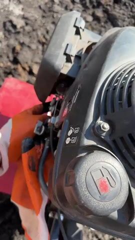 The video begins with a close-up of a person's hands, wearing orange and white work gloves, operating a piece of machinery, possibly a tiller or a similar garden tool. The machine is black with a red fuel cap visible, and some warning labels. The surrounding ground appears to be dark soil.

The scene then shifts to a broader view of an outdoor setting under a bright blue sky with fluffy white clouds. There are some buildings with white siding and darker roofs visible in the distance, along with fences and some greenery. A large white tarp or covering is also present. The sun is shining, suggesting it is daytime. The camera angle is tilted, giving a somewhat distorted perspective of the environment. The overall impression is of a construction site or a rural property being worked on. The location is identified as Rassokha, Russia.