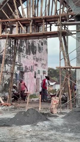 The video shows a construction site with several men working. In the foreground, there are piles of dark grey sand and a red wheelbarrow. A large bag of what appears to be cement is also visible. In the background, a partially constructed building is under scaffolding made of bamboo. Several men are engaged in construction activities, including carrying materials and working with tools. One man, wearing a red shirt and a hat, is pushing a wheelbarrow. Another man, wearing a pink shirt, is sitting down. The building itself appears old and somewhat dilapidated, with peeling pink paint on its walls. Clothes are hanging on a line outside one of the rooms. The overall environment suggests an outdoor construction project, possibly in a rural or developing area, with a clear sky overhead.