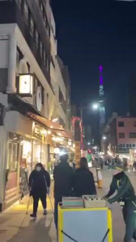 The video shows a nighttime street scene in Japan. The Tokyo Skytree is visible in the distance, illuminated with rainbow colors. The street is lined with shops and restaurants, some of which are lit up. People are walking along the street. There is a man in green work clothes pulling a yellow cart along the street.