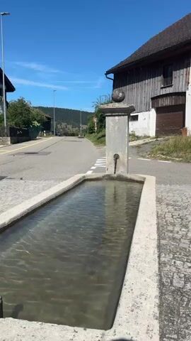 A time-lapse video shows a public water fountain in a rural setting. The fountain is made of concrete and filled with clear water. A faucet is visible on a stone pillar behind the fountain. The background features a paved road, houses, trees, and a hill. The sky is blue and clear. The video captures the subtle movement of the water in the fountain.