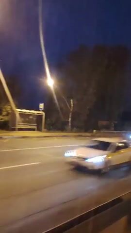 A blurry, low-light video captures a car moving along a road at night. The car, appearing yellow, is in motion, its form blurred by the speed of the vehicle or camera. Streetlights and the dark blue night sky form the background, with some trees visible. A modern-looking building with bright, teal-colored lights is also seen in the distance, suggesting an urban or suburban environment. The perspective suggests the video was taken from another moving vehicle.
