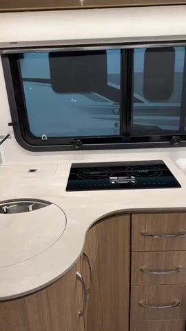 The video shows the interior of a recreational vehicle. The camera moves from the kitchen area, which includes a sink and a stovetop, to a bedroom with two beds, light wood cabinetry and trim, and unique ceiling lighting. The beds are neatly made with plaid comforters.