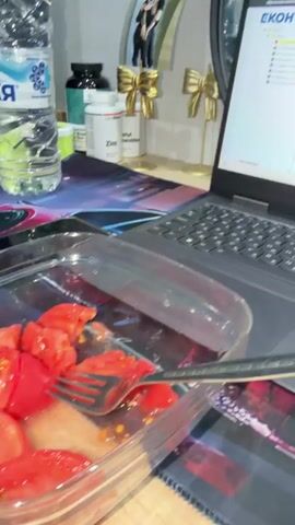 The video shows a close-up of a laptop screen displaying a website with the text "ЕКОНТ" at the top left corner. A laptop is open and positioned on a desk with a colorful mousepad. To the left of the laptop are several bottles, possibly supplements, with labels that are difficult to read. There are also two decorative gold bows in the background, and a framed picture of two people. In front of the laptop, a clear plastic container with a light brown substance is visible, and a silver utensil lies on top of it. A light mint green computer mouse is situated to the right of the container. The lighting suggests an indoor environment, likely a home office or study area. The camera focuses on the desk and its contents, with the laptop screen being a central element. There are no people visibly interacting with the objects in the frame. The overall scene appears to be a still life setup, possibly for an advertisement or product demonstration.