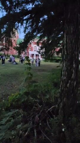 A group of people are gathered in a grassy area in front of a large brick building with a green roof. The building has white trim and multiple windows. The people are standing and sitting, some appear to be talking to each other. There are trees in the foreground and background. The lighting suggests it is either early morning or late evening.