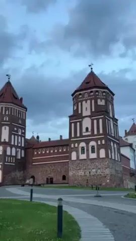 The video shows a view of a historic castle, possibly Mir Castle, from a ground-level perspective. The castle is constructed primarily of red brick and stone. It features multiple towers with red-tiled roofs and white detailing. The sky above is cloudy and dusk seems to be settling. In front of the castle, there is a well-maintained lawn and a paved pathway. Black birds fly in the distance.